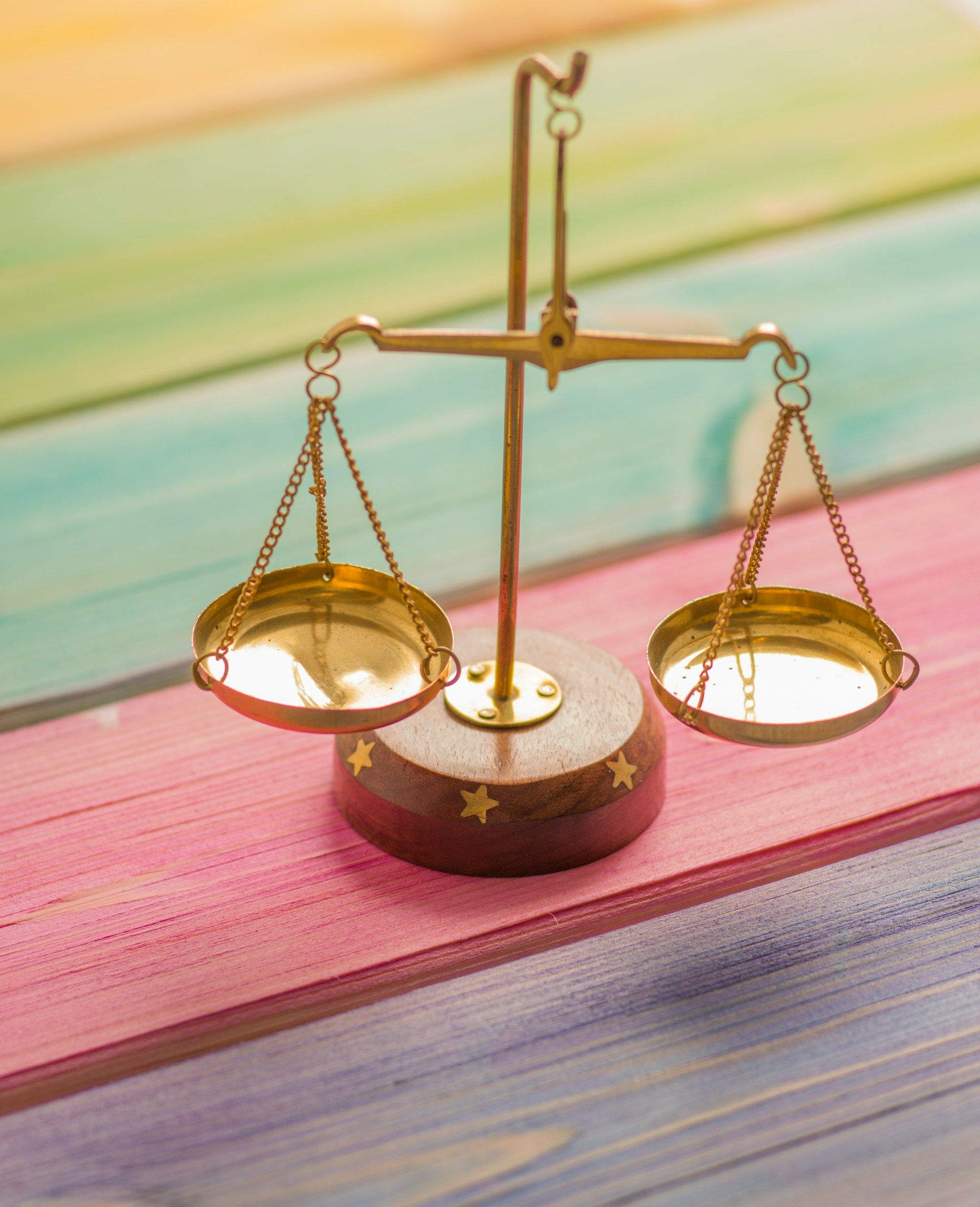 A small, gold-colored balance scale with two hanging pans placed on a colorful wooden surface