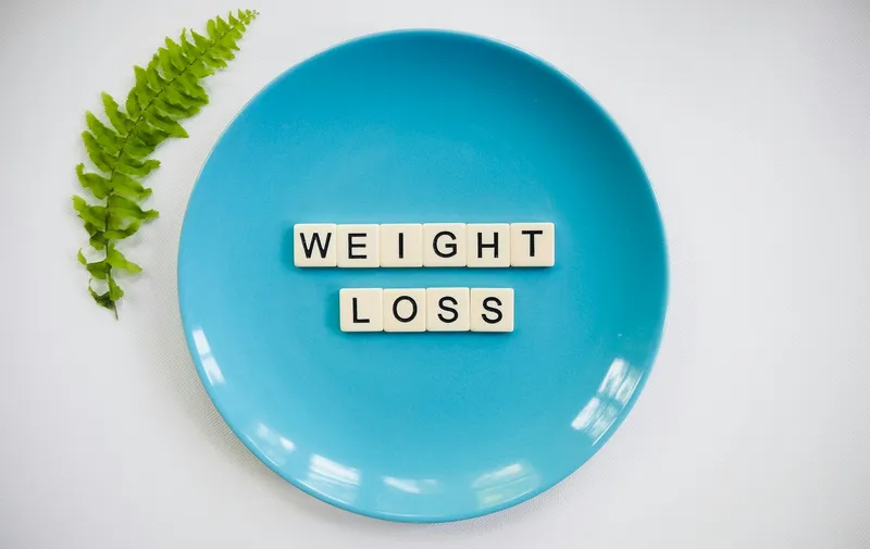 Plato azul con fichas de letras que deletrean “WEIGHT LOSS” y una hoja de helecho verde sobre un fondo blanco.