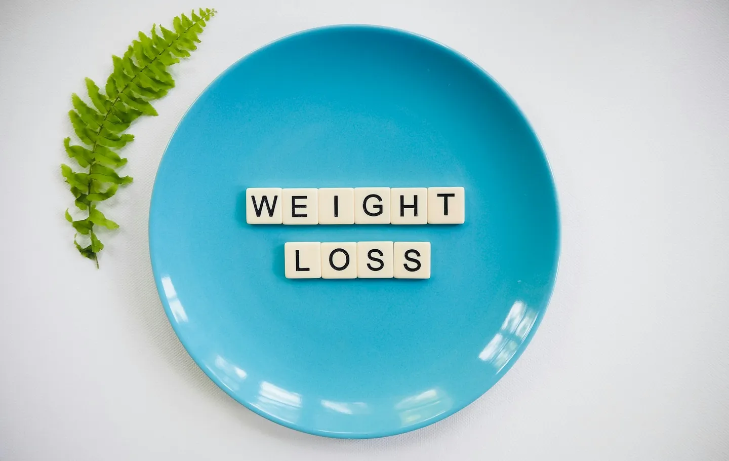Plato azul con fichas de letras que deletrean “WEIGHT LOSS” y una hoja de helecho verde sobre un fondo blanco.