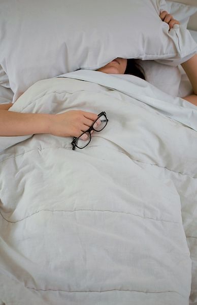 person lying on bed while covering face with pillow while holding eyeglasses
