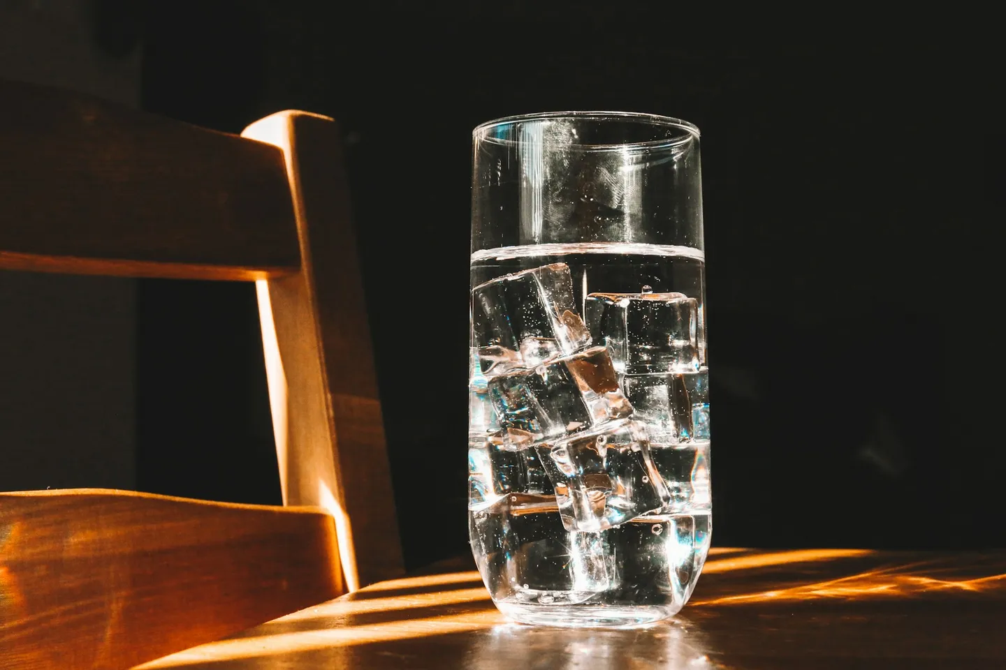 Un vaso alto con agua y hielo sobre una superficie de madera, iluminado por la luz del sol, con un fondo oscuro.