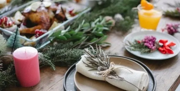 Una mesa festiva navideña con un plato y una servilleta adornados con una ramita verde, rodeados de decoraciones, una vela encendida, un vaso con bebida naranja y platos de comida navideña al fondo.