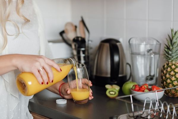 A woman pouring fresh orange juice into a glass in a kitchen surrounded by fruits.